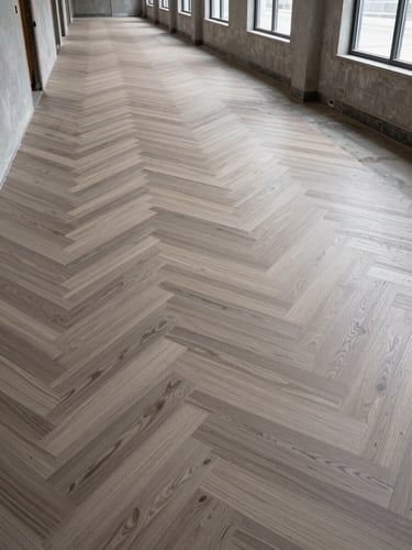 Wide-angle shot down a commercial corridor mid-installation, LVP planks laid in a herringbone pattern across two-thirds of the floor, raw concrete subfloor visible at the far end, overcast daylight from side windows casting even shadowless light, no people present