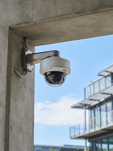 A crisp daylight shot of a modern dome IP security camera mounted on a clean concrete architectural beam, blue sky in the background, professional tech environment.