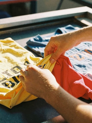 Close-up detail of hands sorting colorful t-shirts in a screen-printing workshop, warm natural light, dust particles in the air, 35mm style