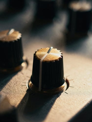 Extreme close-up of an analog synthesizer knob caught in a single sharp beam of sulfur-gold light, deep shadows, dust motes floating in the dark air, cinematic film grain.