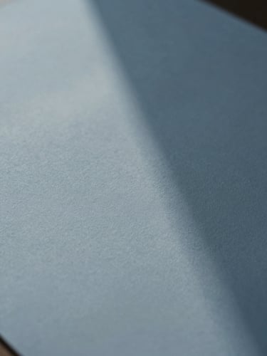 An extreme close-up of fine textured paper casting a soft geometric shadow under diffused daylight, minimalist editorial style, muted blue tones, shallow depth of field.