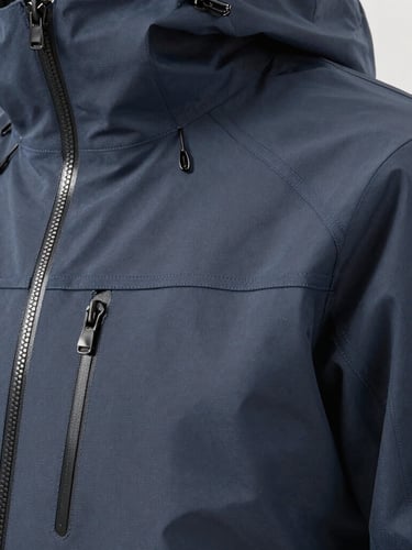 A close-up of a technical waterproof shell jacket showing precise laser-cut pocket edges and heat-sealed zippers, bright clinical lighting, neutral gray background.