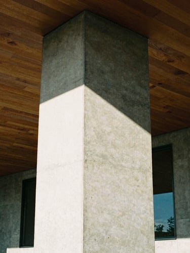 Tight geometric crop of a raw board-formed concrete pillar meeting a high timber ceiling, sharp natural daylight casting deep high-contrast shadows, architectural film photography style.