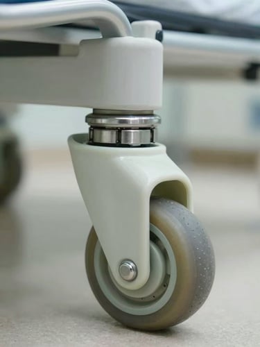 Macro close-up of a heavy-duty hospital bed castor wheel with stainless steel ball bearings and polyurethane tread, bright neutral clinical lighting, sterile environment.