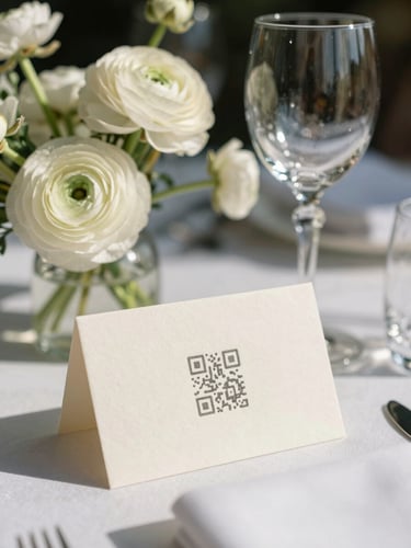 A portrait orientation shot of an elegant wedding tablescape, soft natural daylight, focusing on a beautifully textured ivory place card with a subtle QR code resting next to a crystal wine glass and delicate white ranunculus.