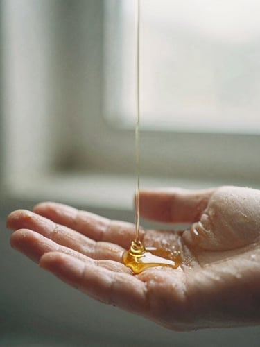 Macro photograph of warm oil dripping onto hands, soft morning window light, muted earth tones, shallow depth of field, 35mm film