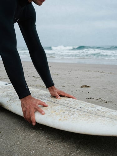 Close-up of a surf instructor's hands adjusting a student's posture on a surfboard on the sand, Playa Cardiel in background, overcast coastal daylight, thick wetsuits, 35mm film grain.