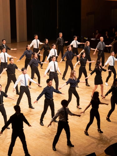 Wide environmental shot of a corporate live-stage performance in progress, rows of performers in synchronised formation under warm golden stage wash, event venue backdrop visible, captured mid-routine with genuine movement blur at the edges