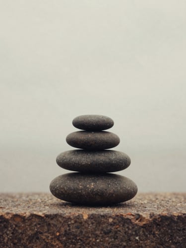 A simple arrangement of three smooth, dark river stones stacked carefully on a warm stone ledge, soft overcast daylight, muted earthy tones, 35mm film style