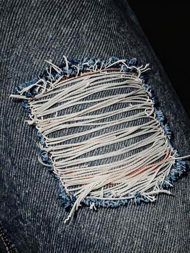 Extreme close-up of frayed denim fabric with heavy physical wear and a faded patch, raw flash photography, desaturated dark tones, high contrast