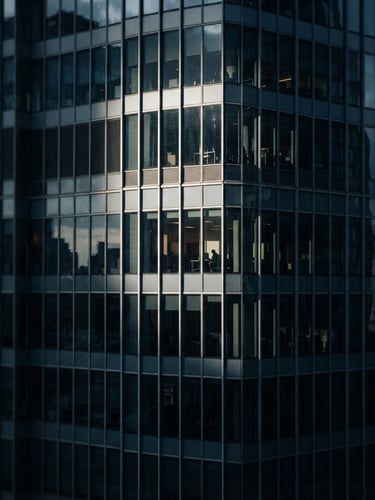 Editorial corporate photography of a modern high-rise glass facade with clean architectural lines, cool slate blue and desaturated navy tones, dramatic window light and deep shadows, shallow depth of field, 35mm.