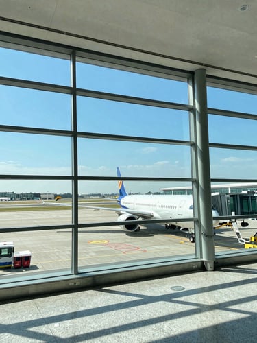 Wide environmental shot of a modern clean airport terminal with bright natural daylight streaming through giant glass windows, a commercial airplane visible on the tarmac
