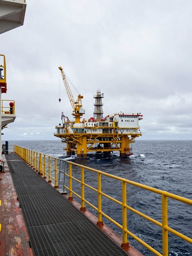 Wide shot from the starboard deck of an offshore production platform at sea, heavy steel grating and safety railings in the foreground, cranes and flare stack visible mid-frame, grey overcast sky and dark North Sea swell extending to the horizon