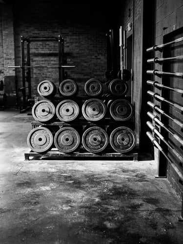 Wide shot of an empty, high-contrast black and white gym floor, heavy iron plates stacked neatly, barbells on racks, harsh side lighting creating deep shadows and bright reflections on metal surfaces, raw, gritty texture.
