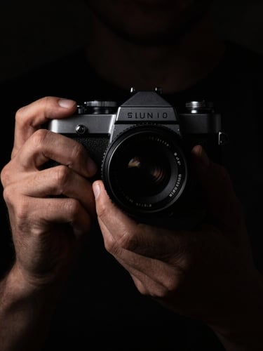 Chiaroscuro portrait of hands holding an old analog camera, deep shadows, single warm light source from the side highlighting the texture of the metallic dials and skin, absolute black background, intimate and quiet framing.