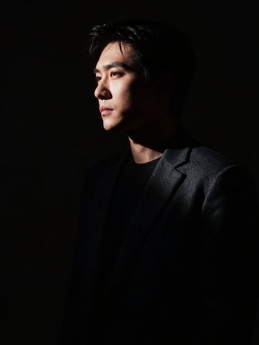 High-contrast chiaroscuro portrait of a solitary figure emerging from deep shadows, single dramatic side light, cinematic framing.