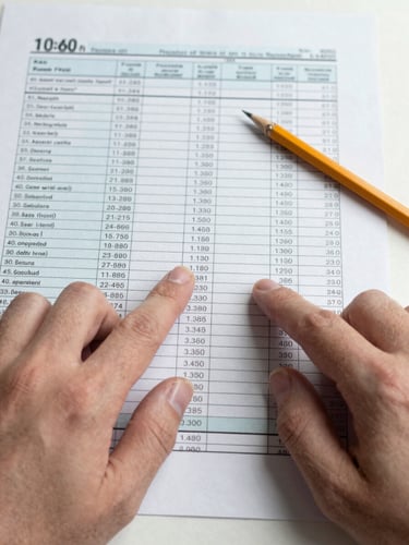 Close-up of hands pointing at tax calculation tables on printed A4 documents, pencil resting beside a column of figures, flat office daylight, no shadows