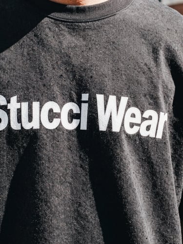 Close-up of a Stucci Wear graphic print on heavyweight cotton, showing fabric texture and stitching detail, harsh direct sunlight, urban background blur