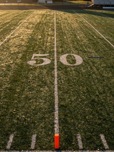 Wide environmental shot looking down the length of a youth flag football field at golden hour, yard lines painted on grass, portable orange pylons at the end zone corners, empty bleachers visible at right edge, long shadow lines stretching across the turf from low sun at left frame