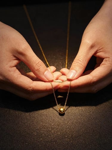 Elegant lifestyle shot focusing on hands gently holding a gold necklace over a dark textured fabric, soft golden reflections, warm directional studio lighting, high contrast