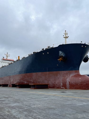 Wide-angle industrial shot of a massive commercial tanker hull resting on keel blocks inside a dry dock, dramatic overcast sky, cool slate and deep navy tones.