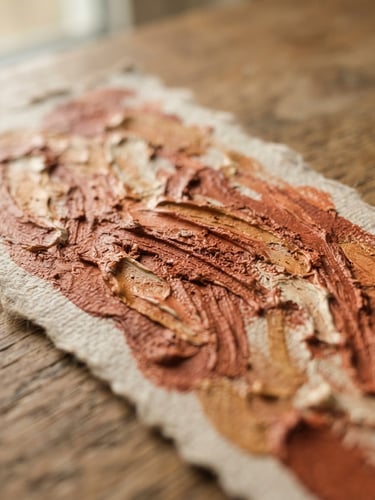 Macro close-up of a hand-painted bookmark on heavyweight canvas paper, natural side-lit window daylight highlighting thick ridges of terracotta acrylic paint, shallow depth of field