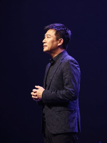 Dramatic profile portrait of a speaker on a dark stage, warm spotlight illuminating their posture, deep navy background, sharp focus, premium editorial photography