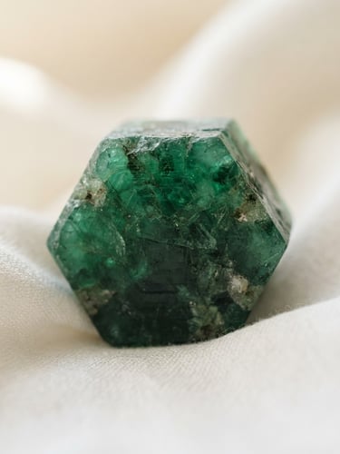 macro editorial photography of a raw, unpolished Colombian emerald crystal resting on clean white silk, soft natural daylight highlighting its deep green untamed inclusions, shallow depth of field, 35mm