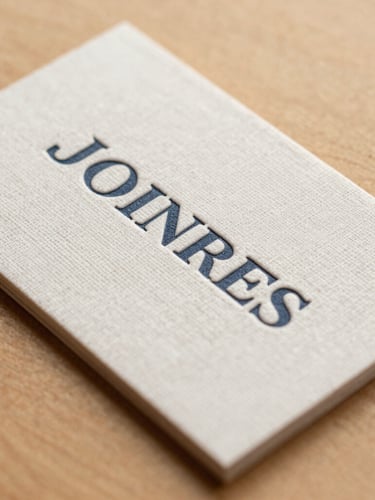 A close-up, high-angle shot of heavily textured linen business cards with embossed typography, resting on a smooth warm almond surface under soft, diffused morning light.