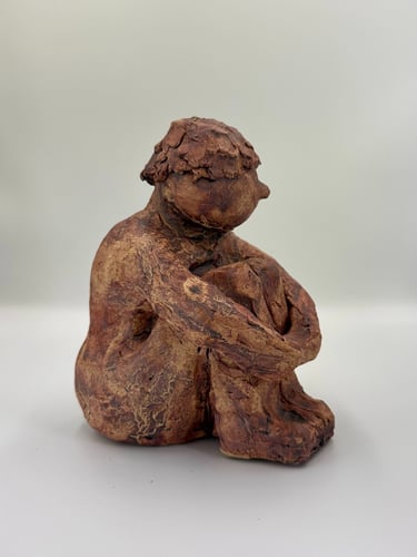 Hand-sculpted reddish-brown clay figurine of a seated person hugging their knees in a pensive pose.
