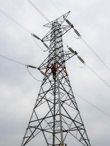 Wide environmental shot from ground level looking up at transmission tower steel lattice, a lineman in rust-red safety harness and hard hat ascending the structure, overcast industrial sky, muted daylight, steel cables converging toward the frame edge, documentary field photography with no studio lighting