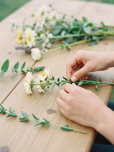 Close-up of hands arranging wildflowers on a rustic wooden table, soft overcast daylight, warm editorial style