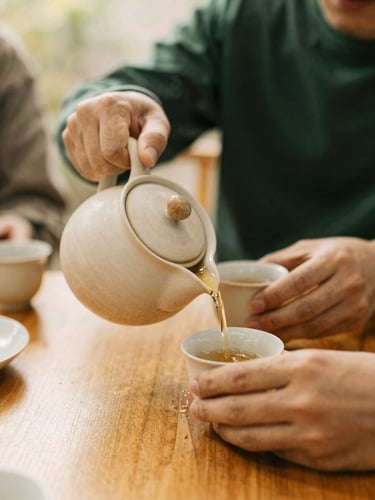 Close-up of hands pouring hot tea from a ceramic pot into cups on a wooden table, warm golden hour light, cozy community atmosphere.