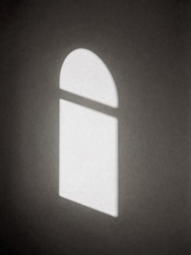 Minimalist black and white photography, soft natural window light casting a long sharp shadow of an arched window onto a bare plaster wall, high contrast, sepia undertones, elegant composition, 35mm film grain