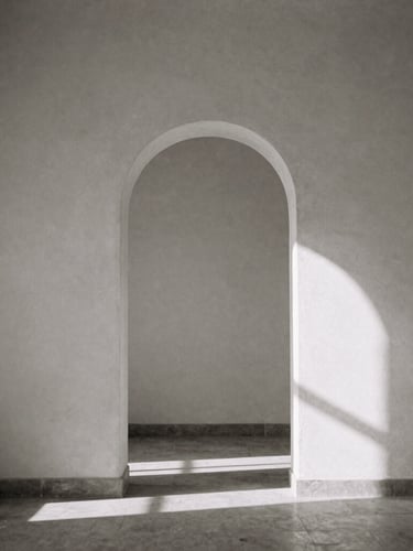 Minimalist black and white photograph with sepia undertones. A solitary stone archway casting a long, geometric shadow on an empty plaster wall, soft directional natural window light, elegant composition, 35mm film grain.