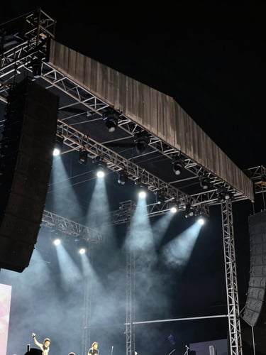 Dramatic portrait shot of a massive concert stage trussing system, heavy line-array speakers suspended, sharp white spotlights cutting through dark haze, high-contrast black background.