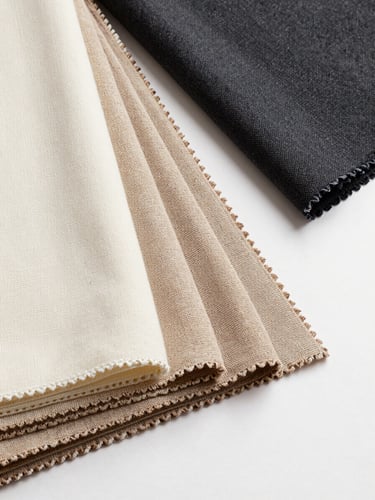 Overhead flat-lay of folded premium woven fabric swatches — ivory, warm beige, and deep charcoal — fanned across a gallery-white surface, close-cropped to reveal weave texture and selvage edge detail, bright controlled studio light casting no shadow