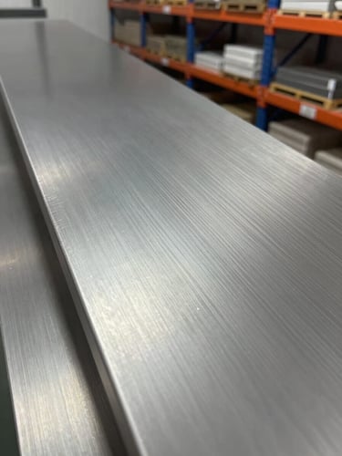 Close-up macro shot of stainless steel sheet surface finish, brushed satin texture visible under cool studio lighting, edge of sheet sharp in left foreground, organized warehouse shelving blurred in background, no people, true metal tone with no color cast
