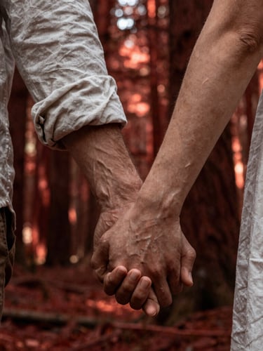 Intimate close-up of a couple holding hands in a deep redwood forest, shafts of dramatic sunlight piercing through the canopy, highlighting creased linen and rich skin tones.