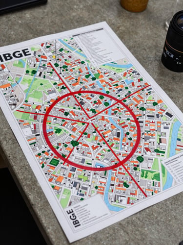 Overhead documentary shot of a Brazilian neighborhood map spread flat on a concrete table, red pen markings circling a flood-risk zone, a printed IBGE data sheet partially visible underneath, flat artificial light from above