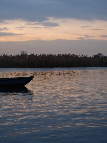 Wide telephoto frame compressed across a Po Delta lagoon at dusk — a flat-bottomed bragozzo silhouetted at the left edge, the water surface layered in deep blue and burnt amber horizontal bands, reed beds fading into soft focus on the far shore, overcast sky bleeding into the water, zero human staging