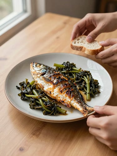 Overhead close-up of a ceramic plate with grilled fish and braised greens on a plain wooden table, hands in frame reaching for bread, natural window light from the left, no styling props