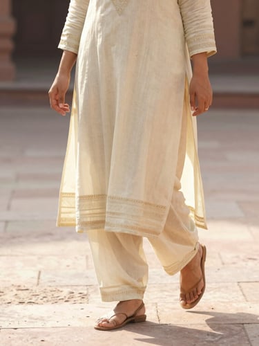 Medium shot of a woman walking gracefully in a cream Farshi salwar suit, showing the elegant drape of wide-legged trousers on a sunlit stone floor, warm editorial style.