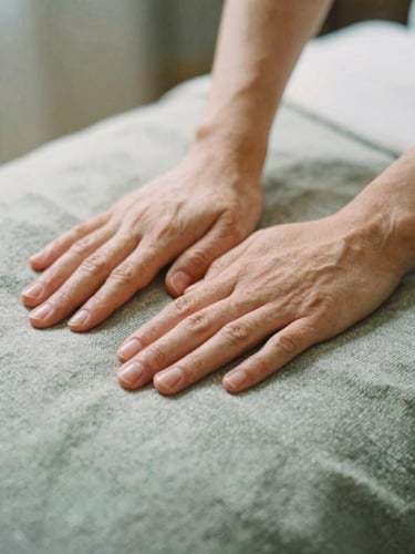 A medium close-up of gentle massage hands resting on a warm, soft textile blanket, soft diffused Nordic daylight, muted sage and stone tones, shallow depth of field, 35mm