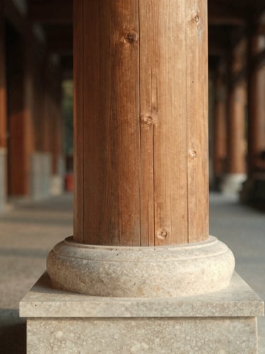 Close-up of a raw timber column meeting a hand-carved travertine base, soft diffused overcast daylight highlighting the coarse textures, deep perspective corridor behind, warm editorial photography.