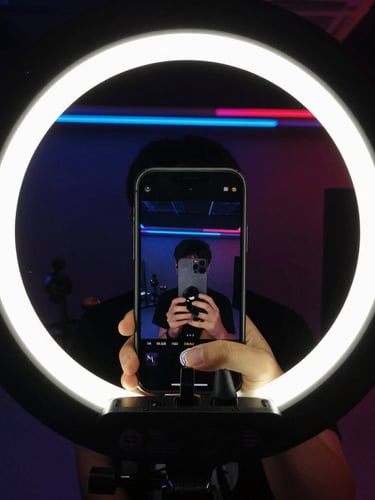 Raw smartphone camera frame with a creator capturing high-contrast behind-the-scenes video under harsh ring light, neon ambient glow.