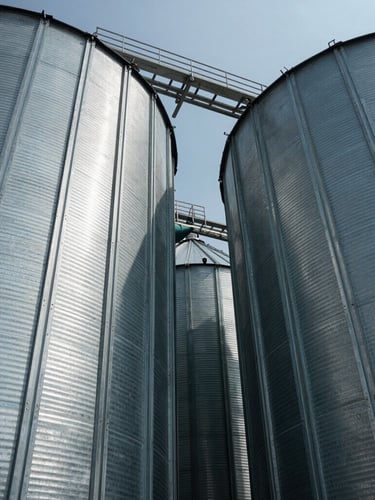 Low-angle shot looking up at massive agricultural silos made of galvanized steel, cold daylight, sharp structural lines, deep shadows.