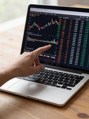 Close-up of a laptop screen on a wooden desk showing real stock market data and index numbers, a hand pointing at the screen, natural daylight from the left, clean and uncluttered
