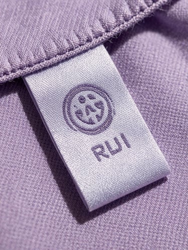 Extreme close-up of a custom-milled organic cotton garment tag stitched with dusty lavender thread, soft natural daylight highlighting fabric texture
