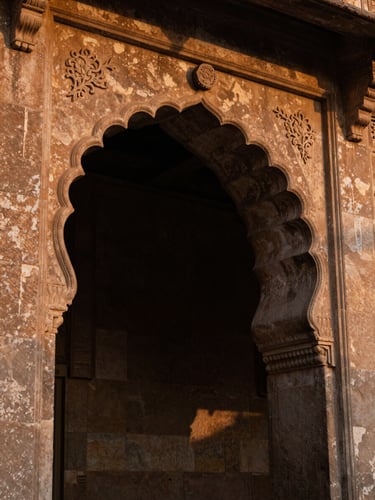 Close-up of weathered Rajasthan stone archway at sidelighting dusk, deep shadows carving the carved floral detail, warm amber and charcoal tones, no people, intimate architectural texture